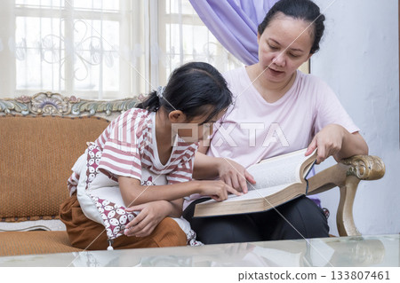 Woman and a child are sitting on a couch reading a book 133807461