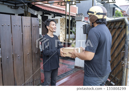 Indonesian southeast asian man receives a cardboard package from a delivery person wearing a helmet. Delivery service 133808299