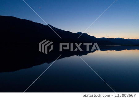 Depicting flat design dusk silhouette of mountain ridge reflecting on calm lake under crescent moon 133808667