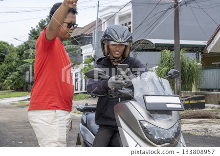 Indonesian southeast asian man wearing a helmet on a motorcycle with a cellphone asking for the location while the other man is pointing and giving directions Indonesian southeast asian man wearing a helmet on a motorcycle with a cellphone asking for the location while the other man is pointing and giving directions 133808957