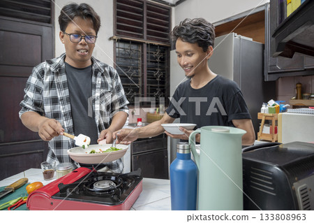 Two Indonesian southeast asian men are cooking together in a kitchen. Cooking in a frying pan on a gas stove, while holding a plate tasting the dish with a spoon. Cooking activity 133808963