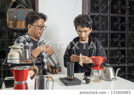Two Indonesian southeast asian men are preparing coffee. Pouring coffee beans into a manual coffee grinder to make coffee at home. Making coffee activity 133808965