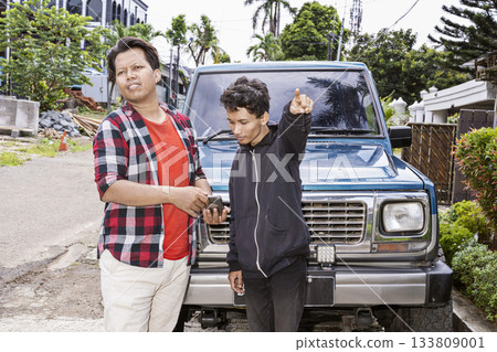Two Indonesian southeast asian men in front of a blue car outside. One man is looking confused and asking the directions while the other is looking at a cellphone and pointing the way 133809001