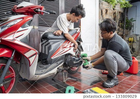 Indonesian southeast asian repairman with a green impact wrench tool discusses replacing the part with his customer after examining a motorcycle in a workshop 133809029