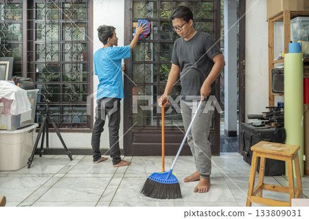 Two Indonesian southeast asian men are cleaning glass windows with a cloth and sweeping the floor with a broom and a dustpan. Cleaning home activity Two Indonesian southeast asian men are cleaning glass windows with a cloth and sweeping the floor with a broom and a dustpan. Cleaning home activity 133809031