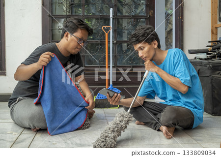 Two Indonesian southeast asian men sitting on the floor while holding cleaning equipment like a rag and a mop relaxed while looking at a cellphone screen. Cleaning home activity Two Indonesian southeast asian men sitting on the floor while holding cleaning equipment like a rag and a mop relaxed while looking at a cellphone screen. Cleaning home activity 133809034