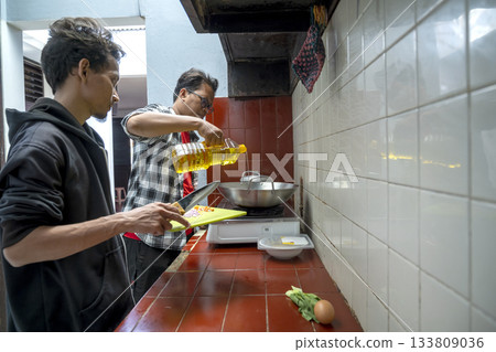 Two Indonesian southeast asian men inside a kitchen while preparing a meal together. Chopped ingredients on a chopping board Two Indonesian southeast asian men inside a kitchen while preparing a meal together. Chopped ingredients on a chopping board 133809036