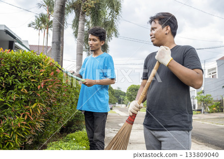 Two Indonesian southeast asian gardeners men trimming a hedge with shears, while the other is sweeping with a broom in the backyard. Gardening work Two Indonesian southeast asian gardeners men trimming a hedge with shears, while the other is sweeping with a broom in the backyard. Gardening work 133809040