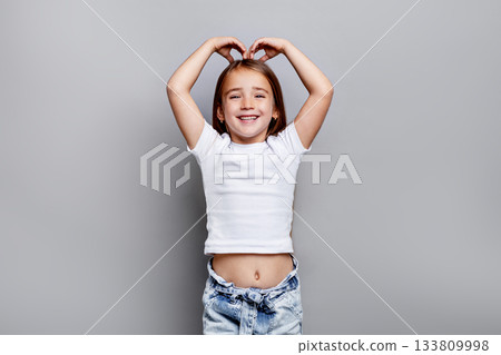 Happy young girl making heart shape with hands above head and smiling in studio, wearing white t-shirt and jeans, joyful portrait against clean grey background Happy young girl making heart shape with hands above head and smiling in studio, wearing white t-shirt and jeans, joyful portrait against clean grey background 133809998