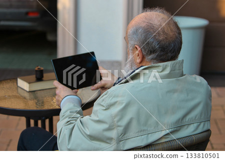Senior man relaxing outdoors with tablet at patio table in peaceful mood 133810501