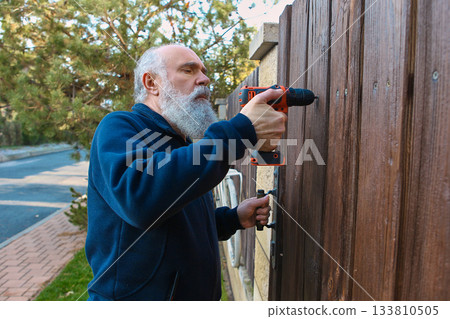 Senior man drilling wooden fence with determined focus during home repair Senior man drilling wooden fence with determined focus during home repair 133810505