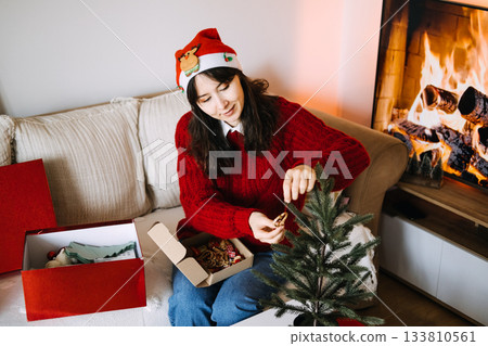 A young woman sits on a sofa decorating a small tabletop Christmas tree with handcrafted straw ornaments. Reusable decor, natural ornaments, sustainable home accents, low-impact Christmas. A young woman sits on a sofa decorating a small tabletop Christmas tree with handcrafted straw ornaments. Reusable decor, natural ornaments, sustainable home accents, low-impact Christmas. 133810561