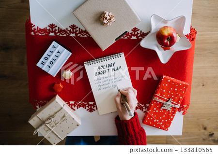 Person writes HOLIDAY BUDGET PLAN in notebook surrounded by wrapped gifts HOLLY JOLLY light box and festive decorations. Holiday budgeting, minimalist gifting, financial wellness, Christmas planning. Person writes HOLIDAY BUDGET PLAN in notebook surrounded by wrapped gifts HOLLY JOLLY light box and festive decorations. Holiday budgeting, minimalist gifting, financial wellness, Christmas planning. 133810565