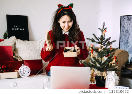Young woman wearing reindeer antlers smiles while holding an open gift box during a video call at home. Digital unboxing, virtual Christmas, remote celebrations, joyful holiday moments Young woman wearing reindeer antlers smiles while holding an open gift box during a video call at home. Digital unboxing, virtual Christmas, remote celebrations, joyful holiday moments 133810632