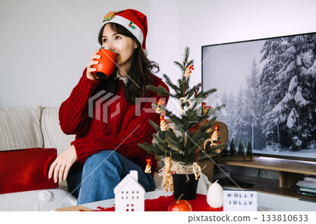 Young woman wearing a Santa hat enjoys a warm drink beside a small Christmas tree while sitting on a couch. Holiday self-care, cozy indoor moments, mindful celebration, winter relaxation Young woman wearing a Santa hat enjoys a warm drink beside a small Christmas tree while sitting on a couch. Holiday self-care, cozy indoor moments, mindful celebration, winter relaxation 133810633