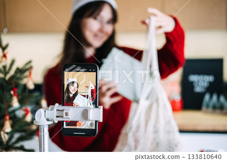 Young adult woman wears Santa hat filming gift unboxing on smartphone using mesh bag in modern kitchen. Eco gifting, sustainable Christmas, zero-waste holiday, ethical unboxing. Young adult woman wears Santa hat filming gift unboxing on smartphone using mesh bag in modern kitchen. Eco gifting, sustainable Christmas, zero-waste holiday, ethical unboxing. 133810640