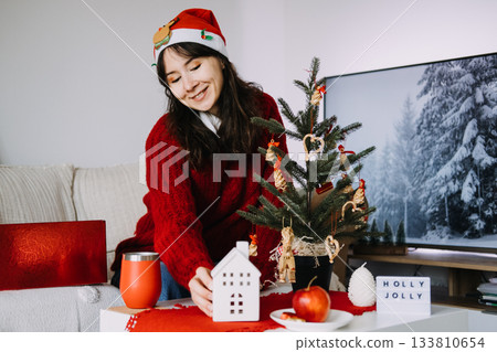 Smiling woman in a Santa hat arranges cozy holiday decorations next to Christmas tree with handmade ornaments. Nostalgic Christmas, handmade ornaments, natural decorations, warm seasonal tradition Smiling woman in a Santa hat arranges cozy holiday decorations next to Christmas tree with handmade ornaments. Nostalgic Christmas, handmade ornaments, natural decorations, warm seasonal tradition 133810654