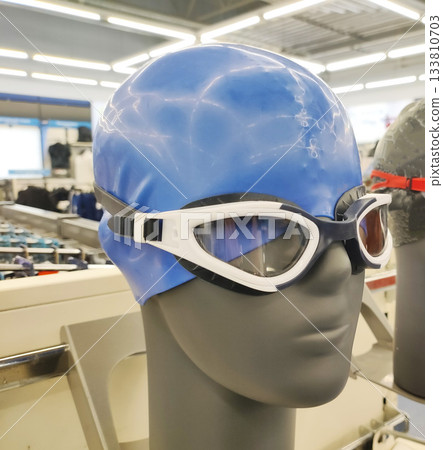 Mannequin head display of blue swimming cap and white swimming goggles in a retail store setting, showcasing essential swimwear accessories for sport and recreation Mannequin head display of blue swimming cap and white swimming goggles in a retail store setting, showcasing essential swimwear accessories for sport and recreation 133810703