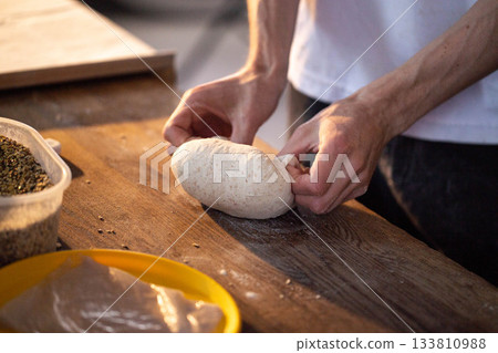 Close up of hands of baker or home cook shaping piece of artisanal bread dough on rustic wooden countertop with natural light highlighting texture of flour and grains 133810988