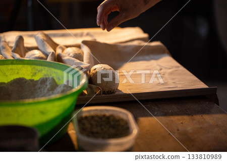Hand of baker preparing dough rolls on baking paper for fresh bread baking, flour in green bowl and spice container visible on rustic wooden table, dark background 133810989