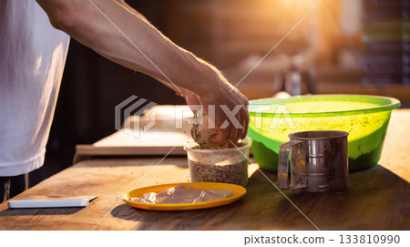 Hands of baker preparing dough, dipping round bread roll in seeds on rustic wooden table, bathed in warm golden light of golden hour, process of artisanal baking creation 133810990