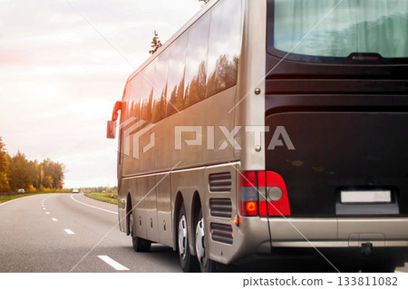 Large coach bus travels on asphalt road during sunset, warm sunlight reflects on vehicle side, trees line roadside, open sky on horizon, view of bus rear. Large coach bus travels on asphalt road during sunset, warm sunlight reflects on vehicle side, trees line roadside, open sky on horizon, view of bus rear. 133811082