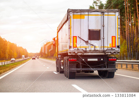 Rear view of white semi-trailer truck with yellow reflective stripes driving on multi-lane asphalt highway during golden hour, sun flare illuminating autumn forest lining roadside, distant vehicles 133811084