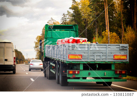 Green semi-truck trailer rear view driving on asphalt highway road through autumn forest landscape under cloudy sky transport cargo vehicle 133811087