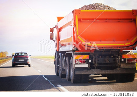 Large orange dump truck carrying gravel on highway with sun flare black car following, transportation of construction materials on open road Large orange dump truck carrying gravel on highway with sun flare black car following, transportation of construction materials on open road 133811088