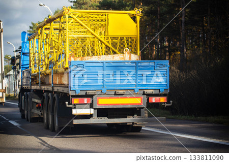 Rear view of long-haul truck trailer with heavy cargo cage on highway at daytime, showcasing industrial transportation and road logistics Rear view of long-haul truck trailer with heavy cargo cage on highway at daytime, showcasing industrial transportation and road logistics 133811090