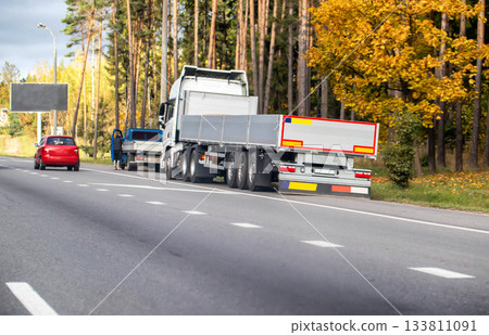 White semi-trailer truck with flatbed trailer parked roadside near red car and person on autumn highway alongside forest with yellow foliage 133811091