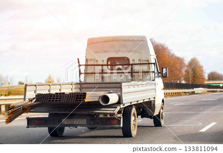White flatbed truck driving on road transporting building materials including metal rebar rods, square profiles, and large pipe 133811094