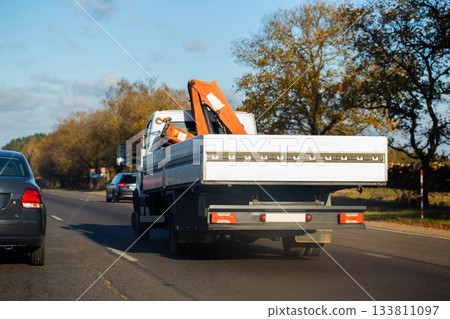 Tow truck with flatbed and crane attachment driving on highway during day, autumn trees lining road, vehicle transportation for heavy loads and recovery service Tow truck with flatbed and crane attachment driving on highway during day, autumn trees lining road, vehicle transportation for heavy loads and recovery service 133811097