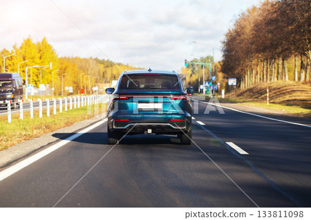 SUV vehicle driving on highway during autumn day. Sunlight illuminating teal car on asphalt road with yellow trees and signs in background, outdoor travel scene SUV vehicle driving on highway during autumn day. Sunlight illuminating teal car on asphalt road with yellow trees and signs in background, outdoor travel scene 133811098
