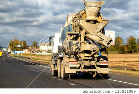 Cement mixer truck driving on asphalt highway road, rear view of vehicle showing large rotating drum and dirty surfaces, transportation of building materials under cloudy autumn sky 133811099