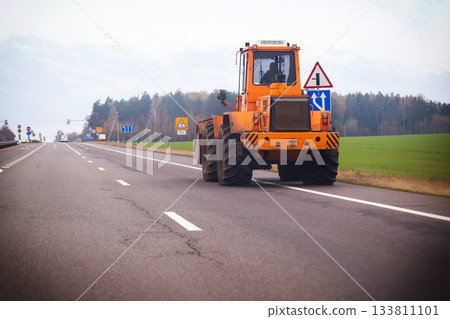 Orange heavy machinery tractor driving away on rural asphalt highway road. Vehicle features large wheels and rear view with tail lights, surrounded by traffic signs, green fields, and distant forest 133811101