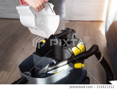 A woman pours clean water and cleaner into a wet dry vacuum cleaner in a room. Concept for cleaning upholstered furniture at home from dust and dirt, professional A woman pours clean water and cleaner into a wet dry vacuum cleaner in a room. Concept for cleaning upholstered furniture at home from dust and dirt, professional 133811312