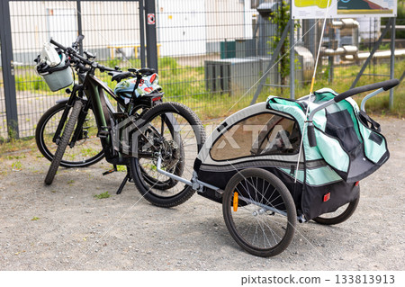 Parked bicycle attached child trailer stands on gravel near industrial fence, showing practical transport setup. Bright colors sturdy construction highlight mobility, family activity outdoor travel Parked bicycle attached child trailer stands on gravel near industrial fence, showing practical transport setup. Bright colors sturdy construction highlight mobility, family activity outdoor travel 133813913