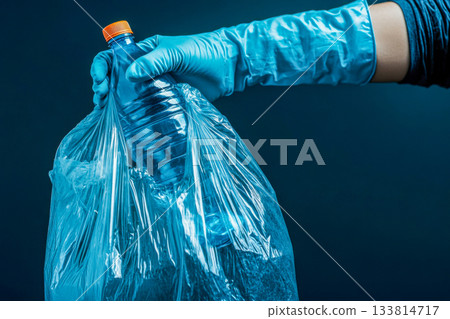 A hand wearing a blue glove holds a plastic bottle inside a clear trash bag, highlighting the importance of recycling and waste management 133814717