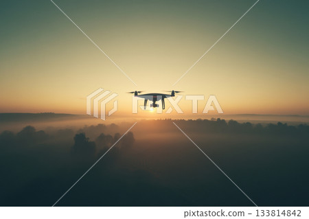 A drone soars above a foggy valley as the sun rises, illuminating the horizon with warm colors in the early morning sky 133814842