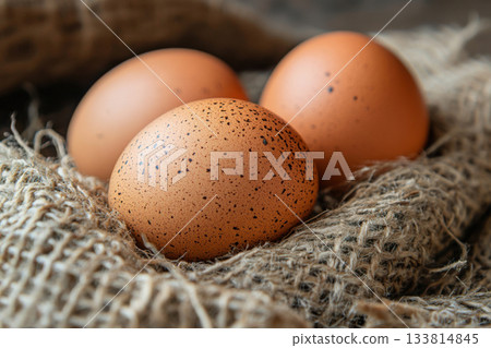 Three brown eggs with speckles are placed on textured burlap fabric, creating a warm and natural kitchen atmosphere 133814845