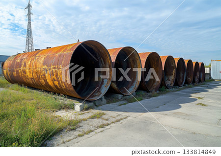 Large, rusted storage tanks are arranged in a row on a concrete path, surrounded by grass and a cloudy sky in the background Large, rusted storage tanks are arranged in a row on a concrete path, surrounded by grass and a cloudy sky in the background 133814849