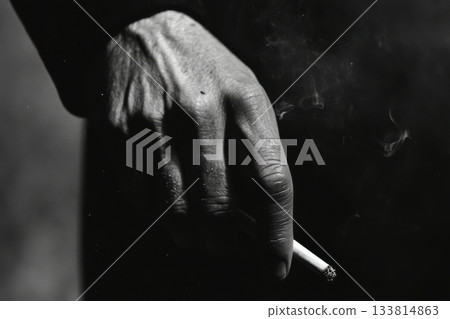 A close-up view shows a hand gripping a cigarette, smoke swirling in the air, capturing a moment of contemplation and solitude 133814863