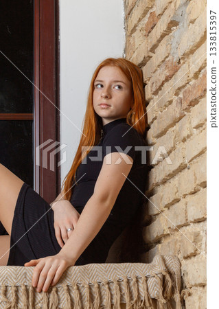 A young red-haired woman sits by a window with a brick wall A young red-haired woman sits by a window with a brick wall 133815397