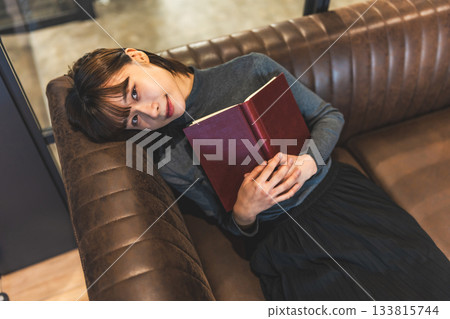 Smiling young woman holding a book on a leather sofa Smiling young woman holding a book on a leather sofa 133815744
