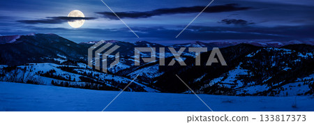 panorama of mountain ridge in winter at night. beautiful landscape with forested hills in snow in full moon light. rural valley in the distance. finance investment or abstract concepts 133817373