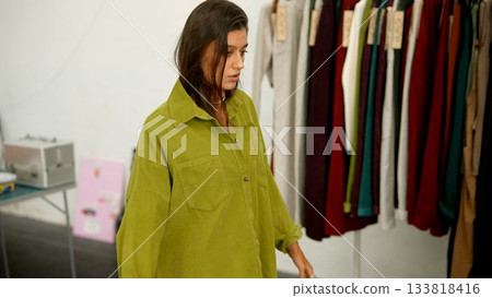 A fashion model is wearing a green oversized shirt while posing next to a clothing rack 133818416