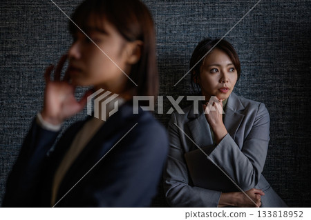 A businesswoman leaning against a wall with a worried expression, and a colleague in front of her looking down 133818952