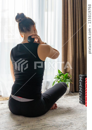 Young woman sitting on the floor at home using a massage ball to relieve neck pain. Self care routine for muscle tension and cervical spine health. 133819768