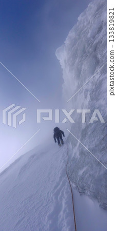 Climber trekking icy mountain ridge through dense fog. Solo mountaineer navigating a challenging snowy ridge against a foggy sky Climber trekking icy mountain ridge through dense fog. Solo mountaineer navigating a challenging snowy ridge against a foggy sky 133819821
